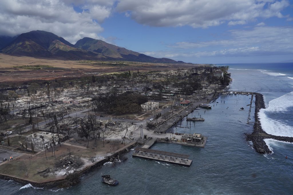 The ruins of Lahania town eerily rests calmly as a large wave breaks over Lahaina Harbor breakwall Thursday, Aug. 10, 2023, in Maui. Two days prior, a large, fast-moving wildfire consumed this historical West Maui town. (Kevin Fujii/Civil Beat/2023)