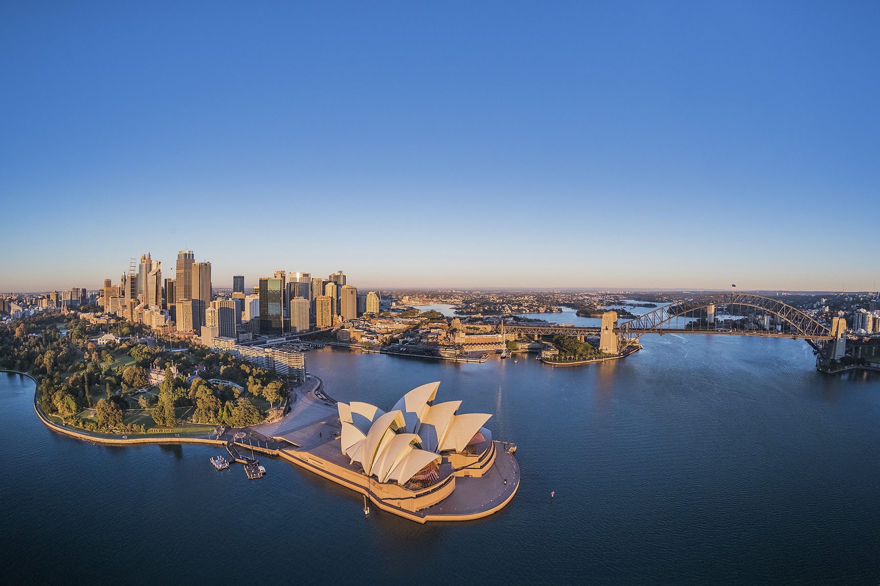 sunset photo of a structure on water that has a roof that points in different directions in the foreground. In the background are a bridge and tall buildings.